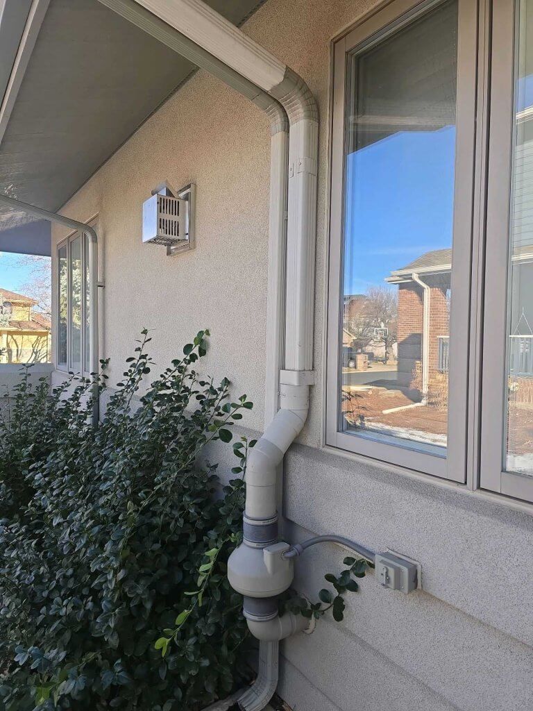 Radon mitigation system on the exterior of a Denver home with white PVC vent pipe extending above the roofline.